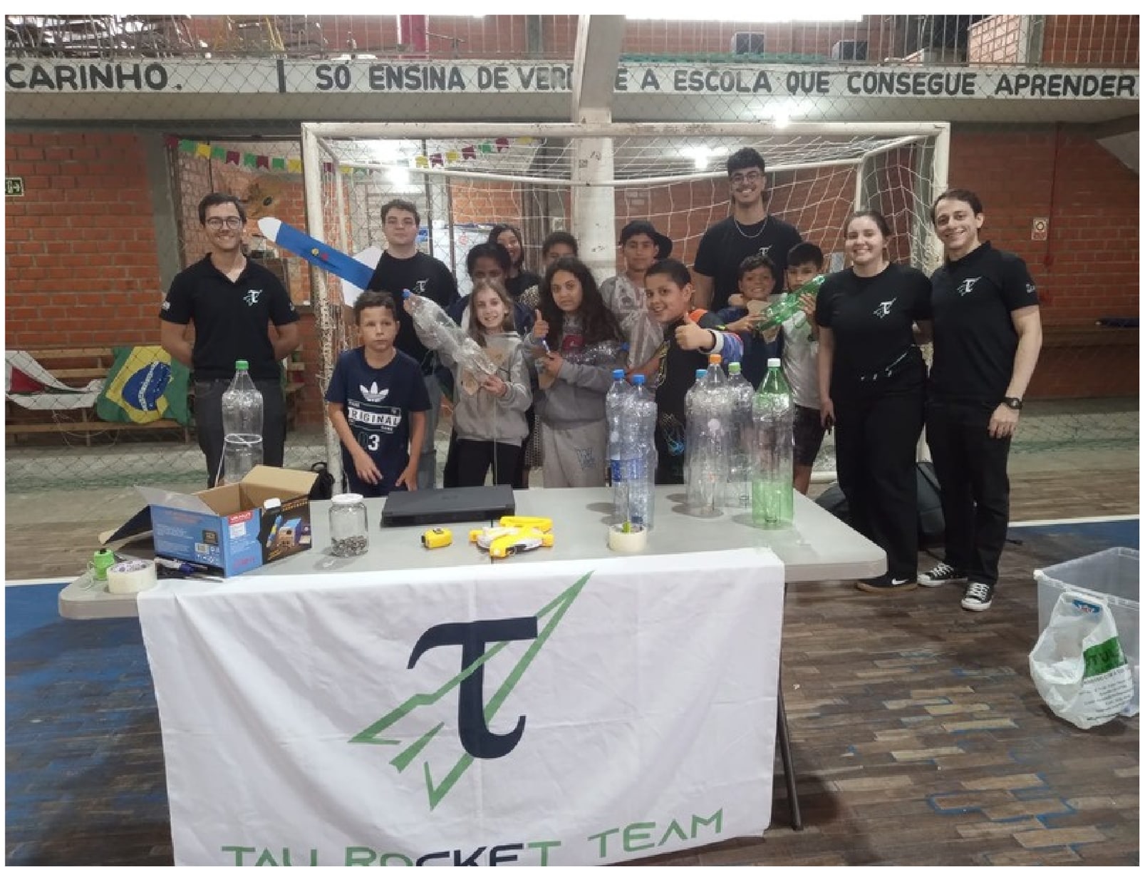 Equipe com crianças em ação de divulgação científica, ao redor da bandeira da Tau Rocket Team